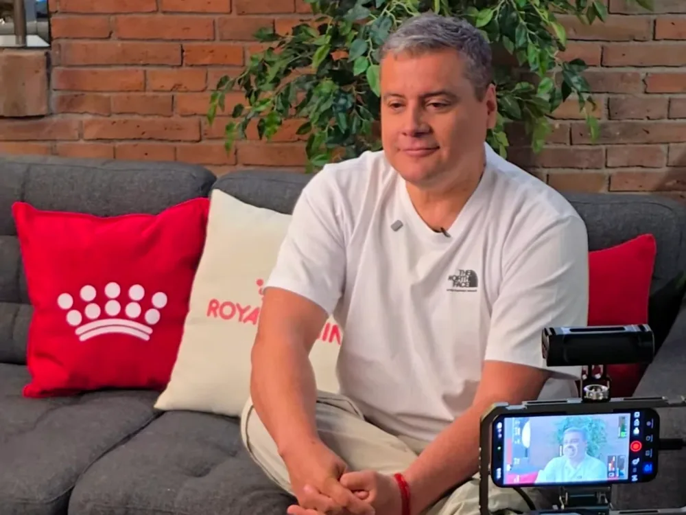 El conductor del programa, sentado en un sofá con almohadones rojos y una pared de ladrillo detrás, durante una entrevista; delante hay un teléfono montado en un trípode que lo graba.