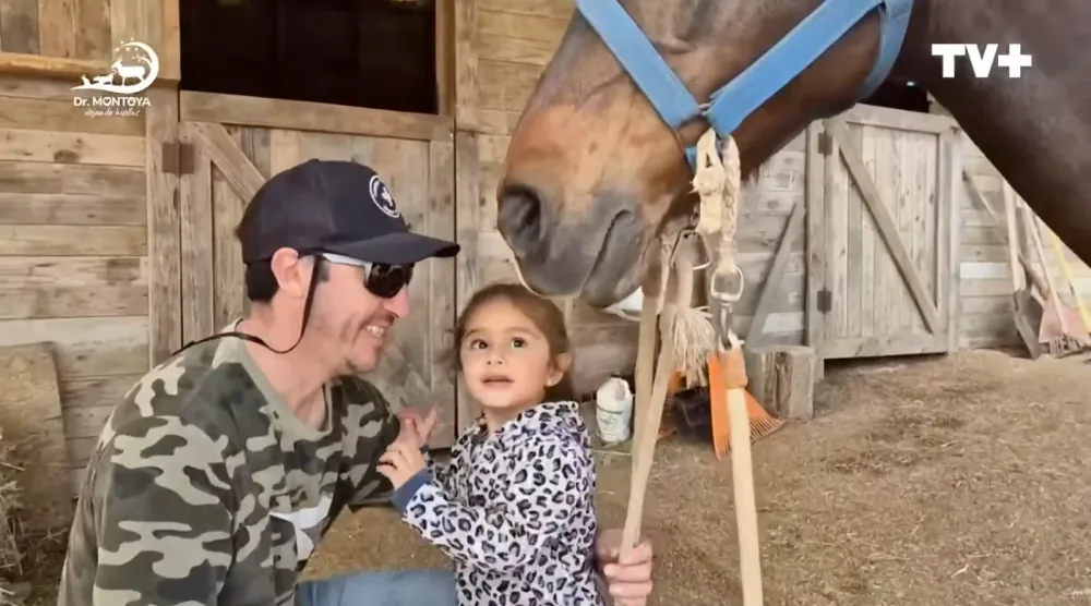 Un hombre con gorra y lentes oscuros se agacha junto a una niña pequeña, ambos sonrientes, frente a un caballo con cabestro azul en un establo de madera.