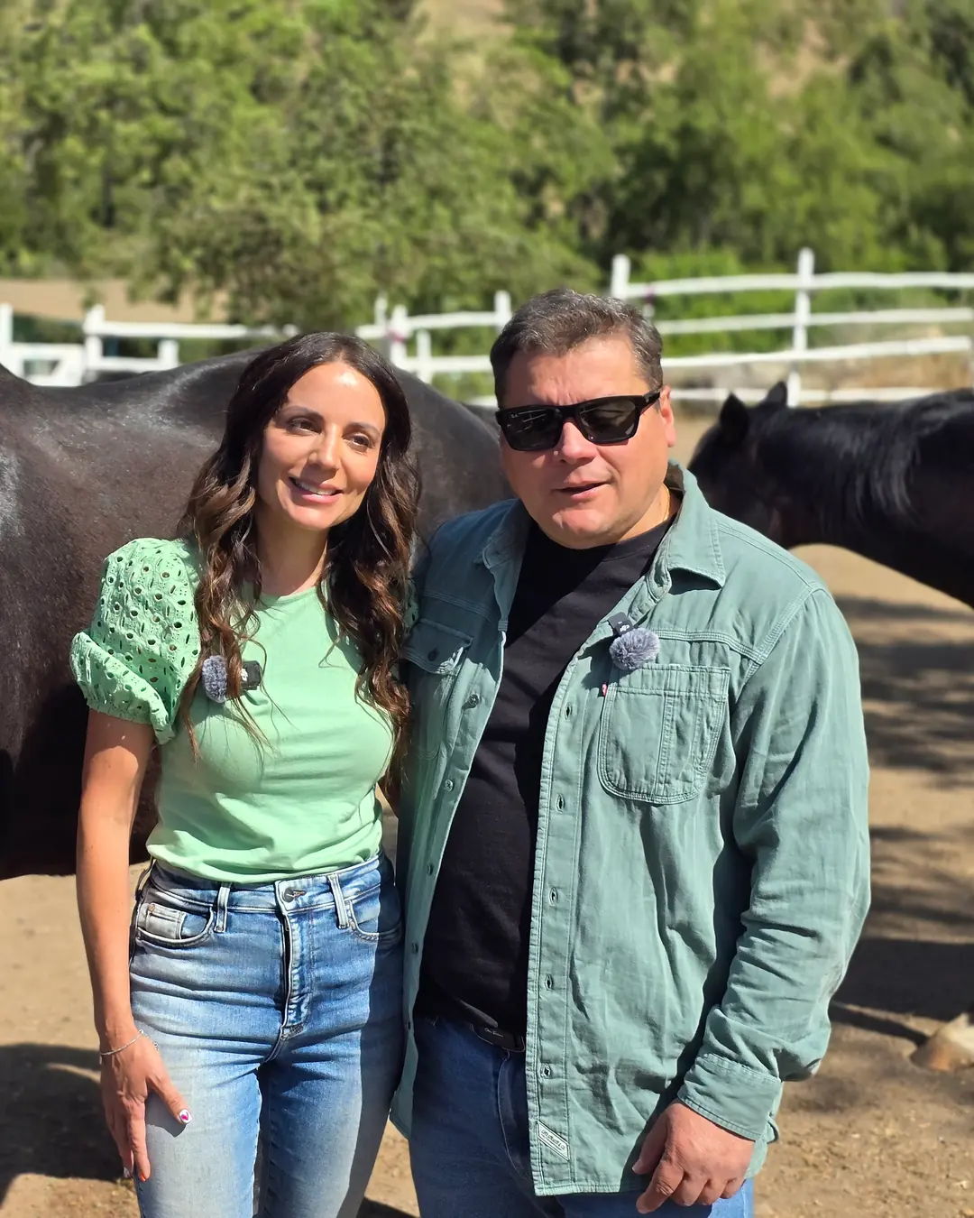 Un hombre con gafas oscuras y una mujer de blusa verde posan junto a un caballo negro con ronzal azul, en un corral con cerca blanca.