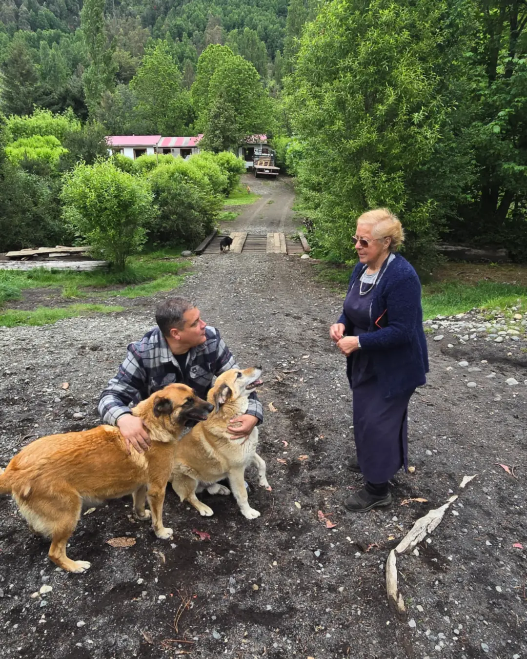 Camino de tierra entre árboles; un hombre agachado acaricia a dos perros dorados mientras una mujer mayor conversa de pie, con un perro negro en primer plano.