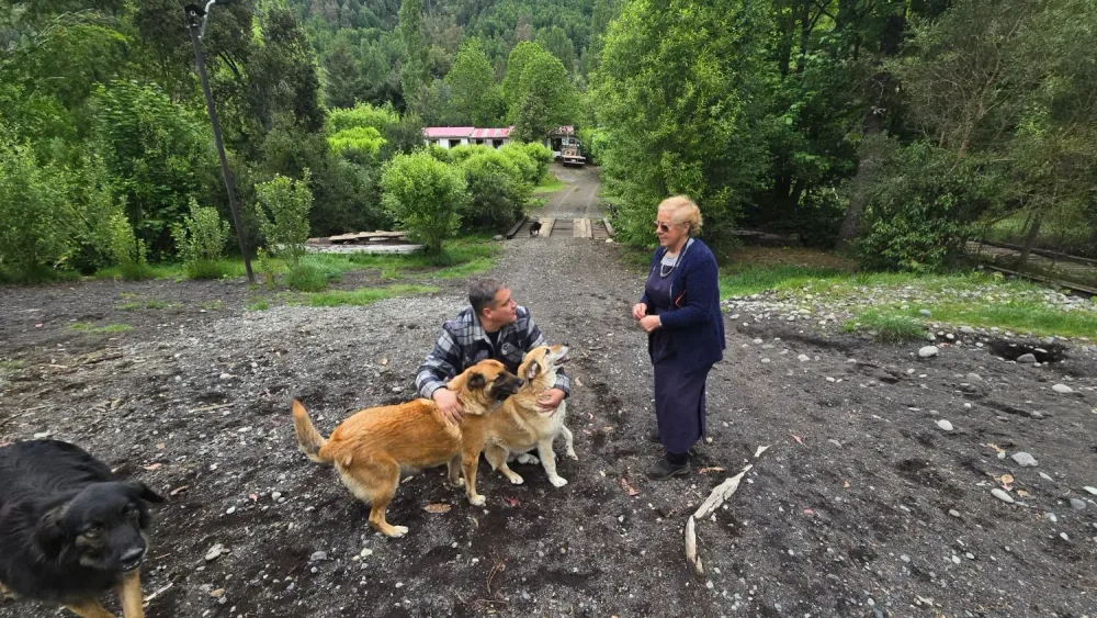 Una mujer mayor de delantal azul conversa con el conductor del programa, que se agacha a acariciar a dos perros color miel, en un camino rodeado de bosque; en primer plano, un perro negro.