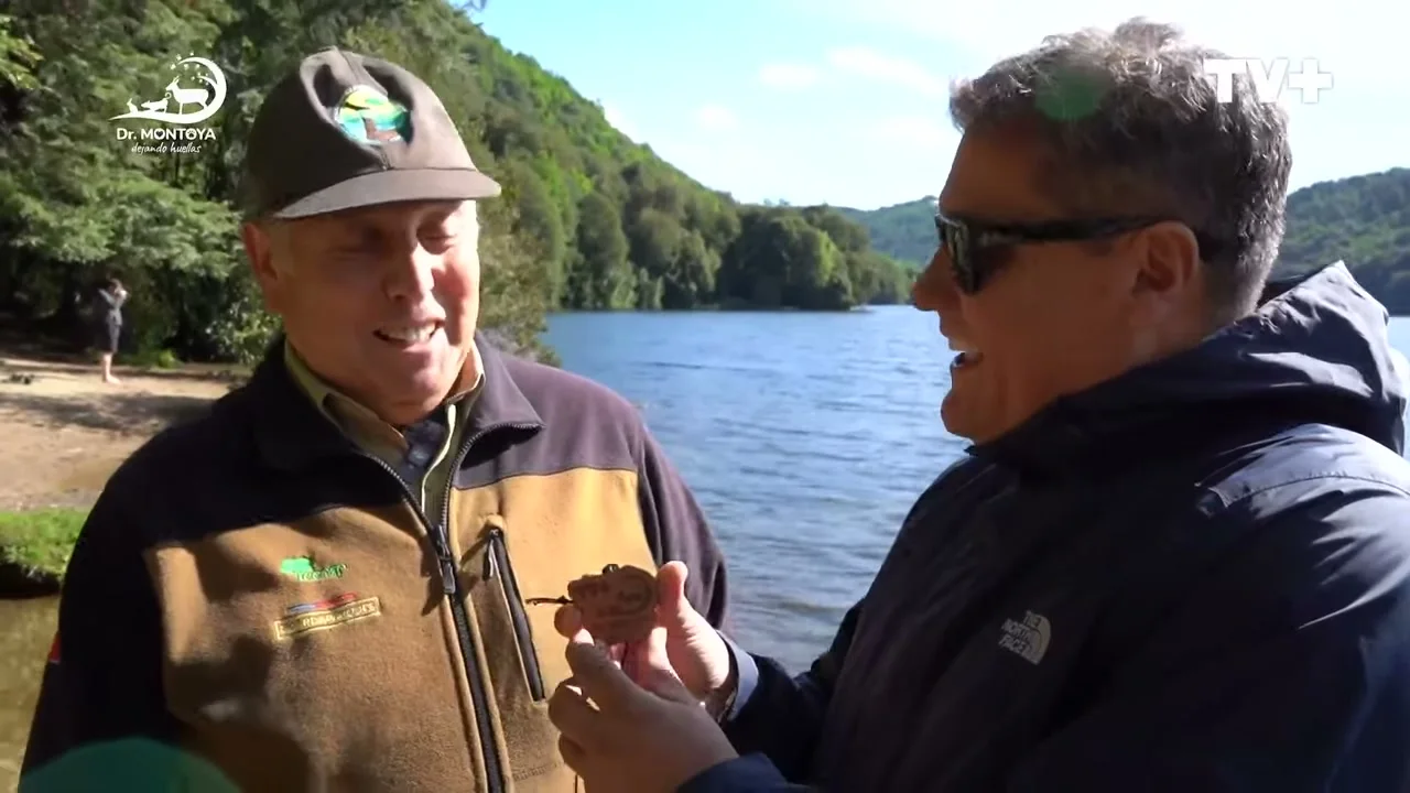 Un guardaparque con gorra y el conductor del programa conversan sonrientes junto a la orilla de un lago rodeado de bosque.